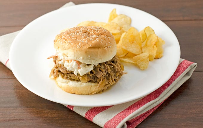 Easy slow cooker pulled pork recipe finished and on a sesame hamburger bun with coleslaw and a side of potato chips on a round white plate sitting on a striped red and white towel on a dark wood tabletop.