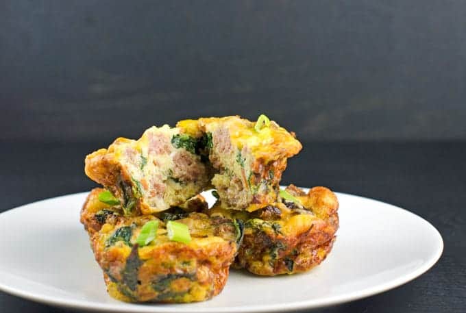 4 baked frittatas with one being cut in half on top sitting on a white plate that is on a black wood background